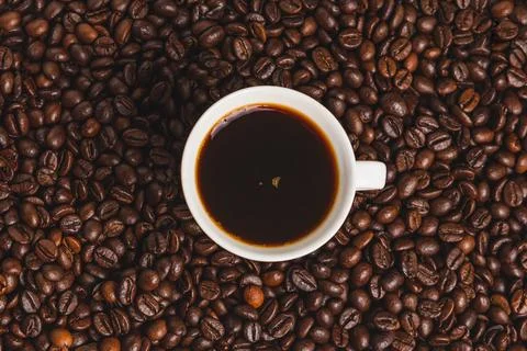 White cup of coffee surrounded by a background of roasted coffee beans. Top.. Stock Photos