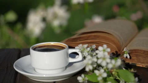 White cup of hot coffee on table with white cherry flowers and book in garden Vidéo 331637155