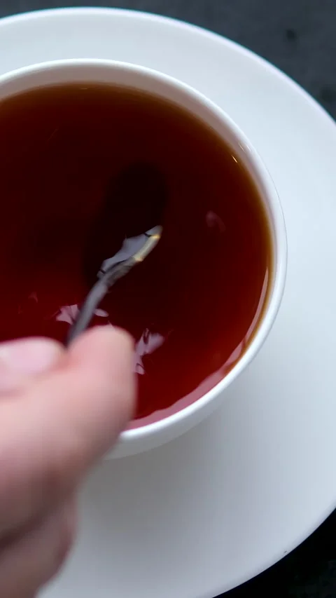 A white cup of tea is being stirred to dissolve the added sugar, resting on a Stock Footage 302066180