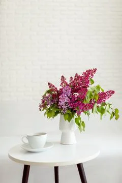 A white cup of tea stands on a table with a bouquet of purple lilacs Stock Photos