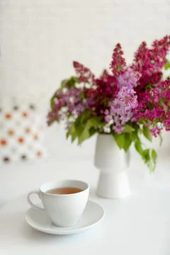 A white cup of tea stands on a table with a bouquet of purple lilacs Stock Photos