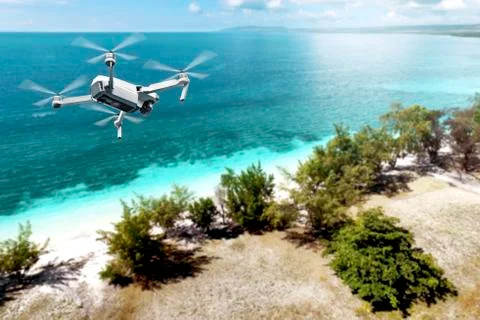 White drone with camera flying over the beach with green trees and blue ocean Stock Photos