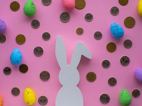 White Easter bunny surrounded by coins and colorful eggs on pink background. Stock Photos