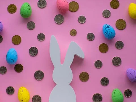 White Easter bunny surrounded by coins and colorful eggs on pink background. Stock Photos