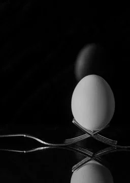 White egg with shell resting on two metal and shiny forks on a black backgrou Stock Photos