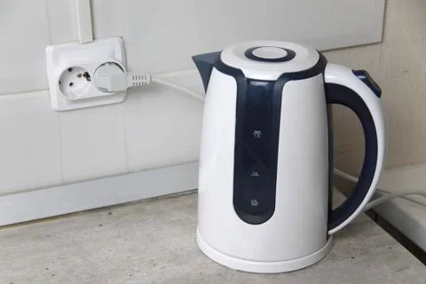 White electric kettle stands on a gray table plugged into a power outlet Stock Photos