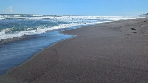 White foam crests over dark volcanic sand as ocean waves approach the shore.. Video stock 331179514