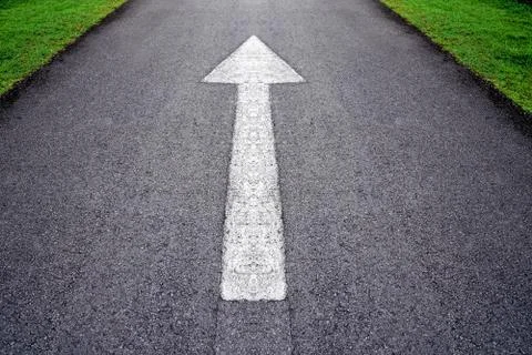 White forward arrow sign on grey asphalt road through the green grass field Stock Photos