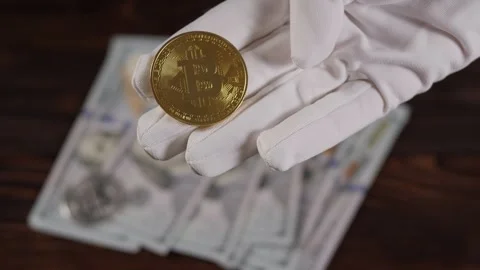 A white-gloved hand holds a bitcoin coin on a background of hundred-dollar bills Stock Footage 279915222