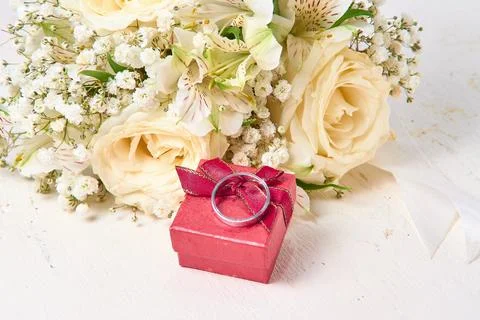 White gold ring rests on red gift box, bouquet of flowers on white textured.. Stock Photos