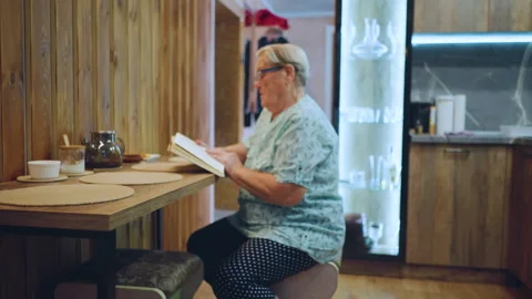 White grandma reading book with tea, wooden table and teapot, floral dress and Stock Footage 334793136
