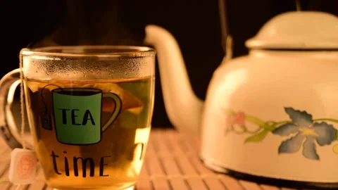 White kettle in the background with cup of tea giving off smoke in front, dar Video stock 154705034