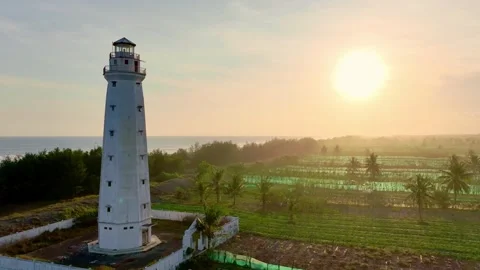 White lighthouse beside sunlit fields and ocean Stock Footage 335433743