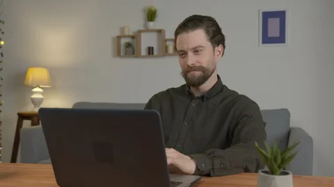 A white man with a beard sits in a room at a table, communicates on the Internet 스톡 동영상 128012105