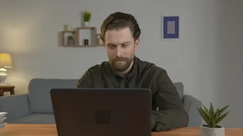 A white man with a beard sits at a table, works on a laptop, works at home Video stock 127390806