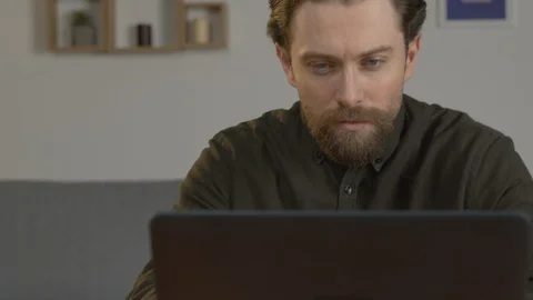 A white man with a beard sits at a table, works on a laptop, works with Stock-Footage 128008614