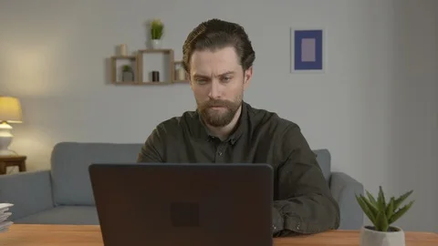 A white man with a beard sits at a table, works on a laptop, works at home Vídeo Stock 128519083