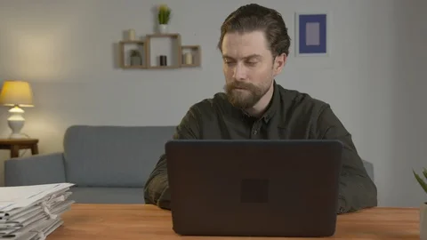 A white man with a beard sits at a table, works on a laptop, works with Vídeo Stock 129074751