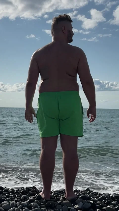 White man with big belly, green shorts on sea pebble beach looks around 库存影片 346896992