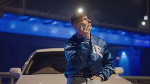 White man leaning on car beneath neon bridge, holding phone to ear, reflective Stock Footage 324952881