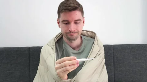 White man sitting on couch wrapped in blanket looking at thermometer Stock Footage 282741227