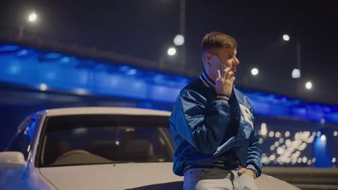 White man talking on phone under bridge leaning on car hood neon lights and Stock Footage 327352920