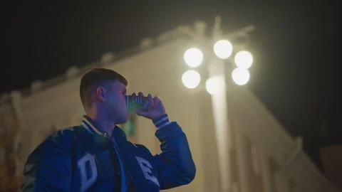 White man under bright stadium lights sipping cold drink in varsity jacket Stock Footage 324957538