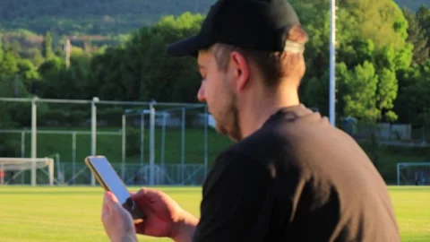 White man using smartphone by football field at sunset with athletes training in Video stock 337555641
