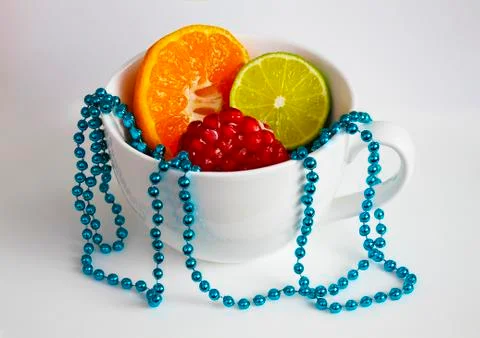 The white mug with cut mandarin, lime, pomegranate and blue beads Stock Photos