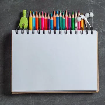 White notebook lies on a set of multi-colored pencils on a dark background Stock Photos