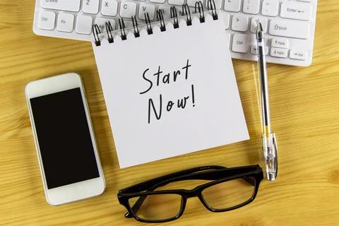 White notepad with text Start Now lying on laptop on wooden table. Stock Photos