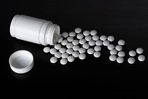 White open jar, scattered tablets, black background. Stock Photos
