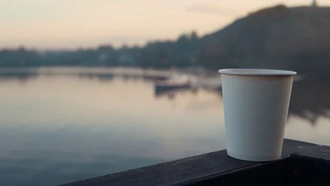 White paper cup with hot coffee stands on the railing above the water at sunset. Stock Footage 96154621