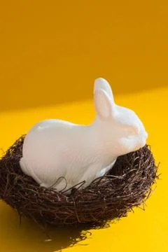 White plastic easter bunny sitting in a nest against a bright yellow background Stock Photos