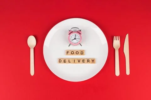 White plate, pink alarm clock, wooden cutlery set and words FOOD DELIVERY on Stock Photos