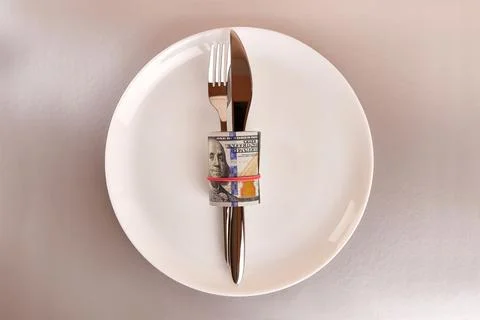 A white plate with a roll of dollars, a fork and a knife on a gray background Stock Photos