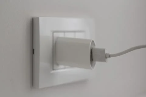White plug from device is plugged into light socket on beige gray wall of hotel Stock Photos