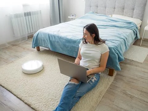 White robotic vacuum cleaner cleaning the floor while woman using laptop Stock Photos