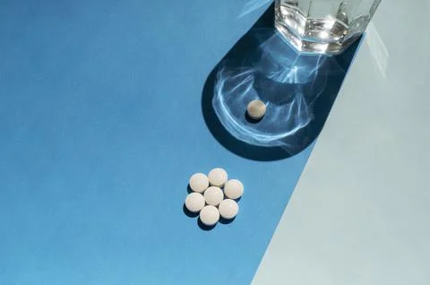 White round pills with one separate tablet and a glass of water on a blue Stock Photos