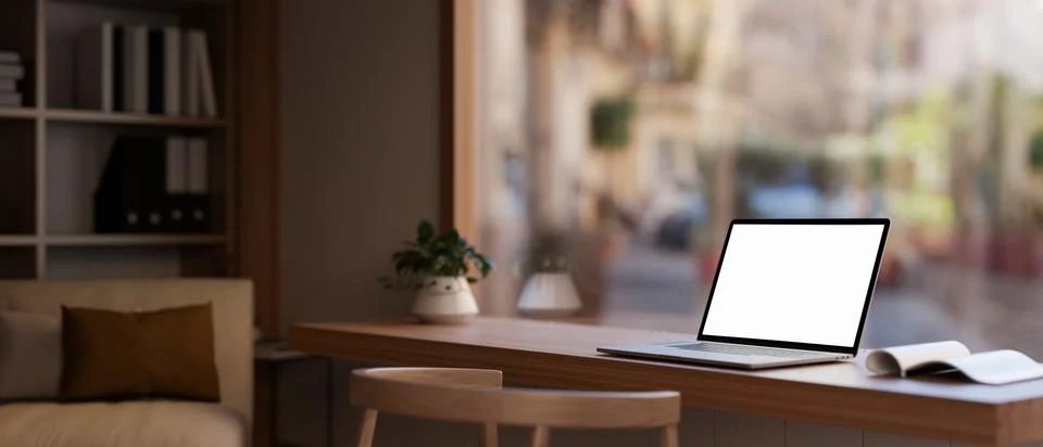 A white-screen laptop computer on a table near a large glass window in a mo.. Illustrazione stock