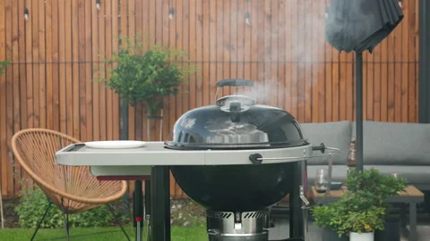 White smoke rises from a closed outdoor charcoal grill on a backyard patio 库存影片 340164391