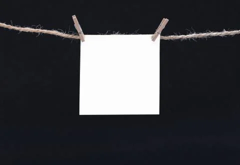 A white square piece of note paper hangs on clothespins on a black background Stock Photos
