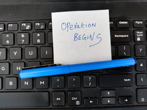 A White sticky note with text OPERATION BEGINS and pen on top of laptop keybo Stock Photos