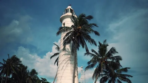 A white stone lighthouse surrounded by palm trees against a blue sky background Stock Footage 267698512