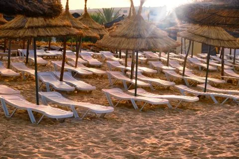White sunbeds on the evening empty beach under straw umbrellas. Sunset sun on Stock Photos