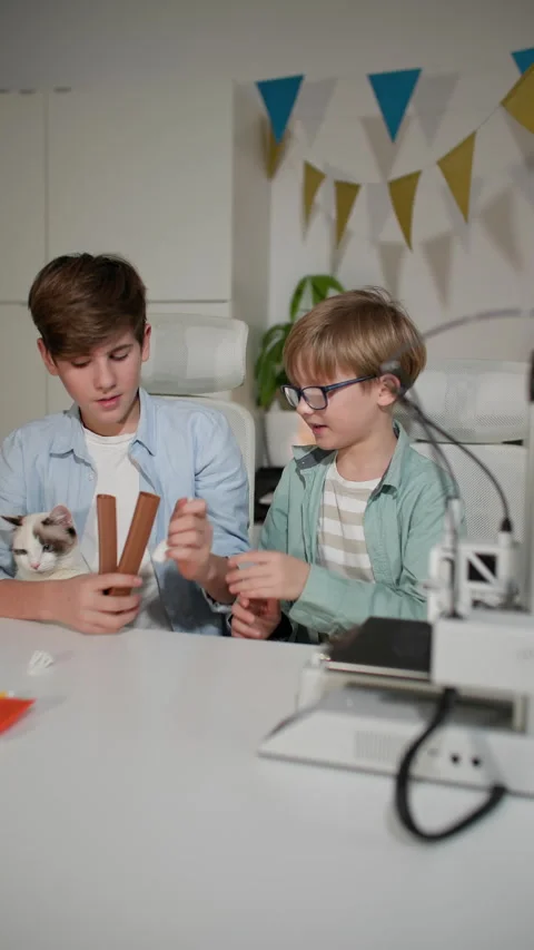 At a white table, two children and a kitten watch a 3D printer in operation Stock Footage 335390019