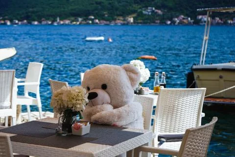 A white teddy bear is sitting at a cafe table on the shore of a lake, river, sea Stock Photos