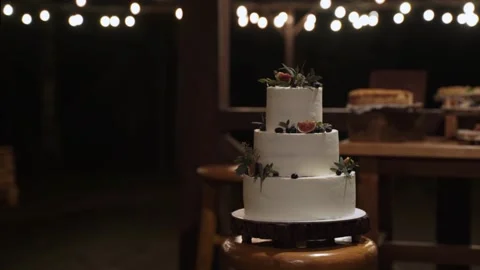 White three-tiered cake, decorated with berries stands outside on wooden tray. Stock Footage 168598715