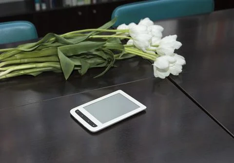 White tulips and e-reader on dark table with bookshelves in background Stock Photos