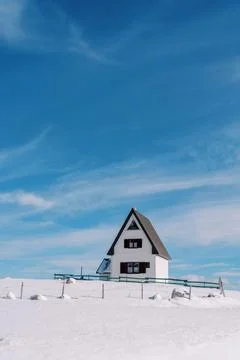 White two-story house with a triangular roof with a wooden fence on a snowy hill Stock Photos